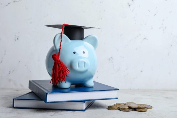 An image of a blue piggy bank with a graduation cap on top of 2 books piled on a table beside coins