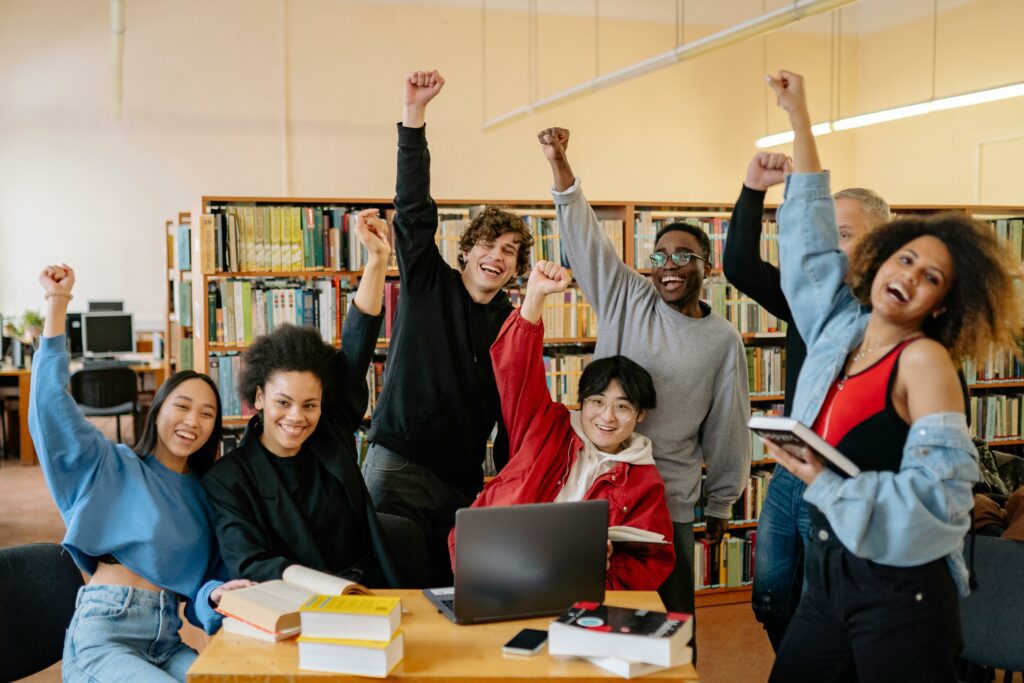 An image of a group of students with different nationalities raising their hands in victory with smiles on their faces inside the library.